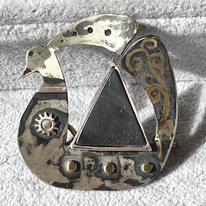 Op de afbeelding: Zilverkleurige metalen broche met een vogelachtig ontwerp, een donkere driehoek en gouden accenten. De broche heeft een zonachtig embleem en drie gouden klinknagels. Het ontwerp doet denken aan Vikingkunst.