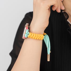 May include: A vibrant Apple Watch strap with a braided yellow and turquoise design. The strap is accented with pink and gold details. The watch strap is shown on a wrist.