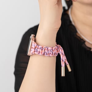 Op de afbeelding: Een roze en paarse gevlochten Apple Watch band met goudkleurige metalen accenten. De band is gemaakt van een geweven materiaal en wordt vastgezet met een lus- en knoopsluiting. De wijzerplaat van de horloge is roségoud.