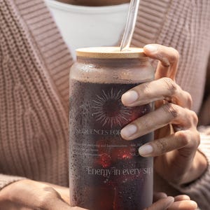 May include: A clear glass can-shaped tumbler filled with a dark red liquid, featuring a bamboo lid and a clear glass straw. The glass has text and a sunburst design. The words "Energy in every sip" are visible.