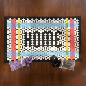 May include: A rectangular doormat with a black border and a honeycomb pattern. The word "HOME" is spelled out in white hexagons. The mat has a rainbow-colored border and comes with bags of purple and black game pieces.