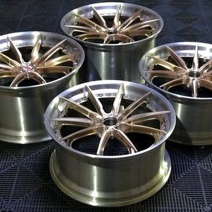 May include: Four alloy car wheels with a brushed silver finish and bronze-coloured spokes. The wheels have a multi-spoke design and a polished appearance. The wheels are arranged in a square formation, showcasing their design and finish.