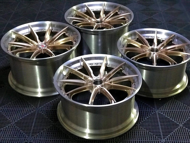 May include: Four alloy car wheels with a brushed silver finish and bronze-coloured spokes. The wheels have a multi-spoke design and a polished appearance. The wheels are arranged in a square formation, showcasing their design and finish.
