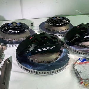 Può includere: Un assortimento di pinze freno e rotori SRT neri, con dettagli argento e blu, disposti su una superficie bianca. Sono incluse anche bottiglie di liquido dei freni, staffe e tubi freno confezionati. Componenti automobilistici.