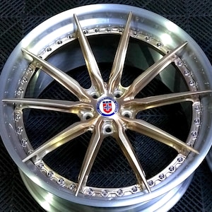 May include: A close-up of a custom car wheel with a gold-coloured spoke design. The wheel has a silver rim with small bolts around the edge. The centre cap features a logo with red, white, and blue colours.