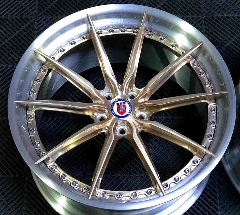 May include: A close-up of a custom car wheel with a gold-coloured spoke design. The wheel has a silver rim with small bolts around the edge. The centre cap features a logo with red, white, and blue colours.
