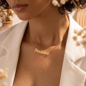 Peut inclure: Un collier doré avec le nom "Madeline" écrit en cursive. Le collier est porté sur une veste blanche. L'image est un gros plan, mettant l'accent sur le collier et le haut du corps.