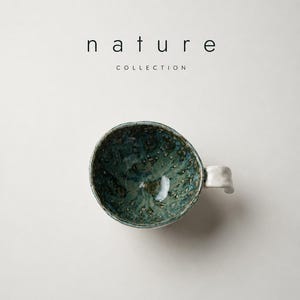 May include: A ceramic mug with a textured, mottled green and blue interior glaze and a white exterior handle. The text "nature COLLECTION" is printed above the mug in black, sans-serif lettering. The mug has an irregular, organic rim shape.