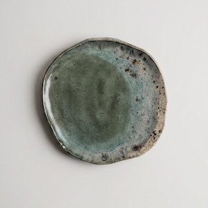 Peut inclure: Une assiette en céramique faite à la main, de forme organique et irrégulière. L'assiette présente un mélange de teintes turquoise et vertes avec une surface tachetée et texturée. Le bord a un bord contrastant de couleur claire.