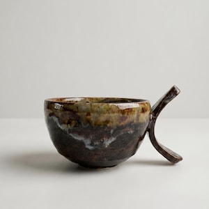 Peut inclure: Une tasse en céramique avec une forme de bol arrondie et une poignée angulaire unique s'étendant vers l'extérieur. La surface présente un émail tacheté aux tons brun profond, gris anthracite foncé et vert olive terreux.