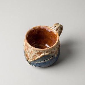 Peut inclure: Mug en céramique fait à la main avec un design unique. L'intérieur est brun foncé, tandis que l'extérieur présente un mélange de bleu, beige et marron. La tasse a une anse robuste et une surface texturée, idéale pour les boissons chaudes.