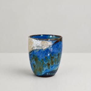 May include: A ceramic cup featuring a reactive glaze with deep blue, cobalt, and earthy brown tones. The upper rim displays a speckled cream and white section that transitions into a vibrant blue body with dark brown, vertical drip-like patterns.