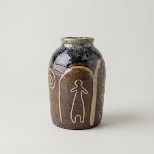 May include: A brown ceramic vase with a dark blue and black rim. The vase features cream-coloured line drawings of figures and spiral designs. The vase is approximately 18 cm tall and has a rounded shape.