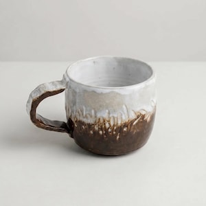 May include: A handcrafted ceramic mug with a distinctive design. The mug has a white interior, fading to light brown and a dark brown base. The handle is textured, giving the mug a rustic, artisanal appearance.