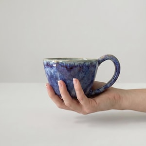 以下が含まれることがあります： 幅の広い丸みを帯びたボウル型の陶器製マグカップで、曲線的なハンドルが付いています。外側は青、紫、白の深い色合いが混ざり合った釉薬で仕上げられており、表面に質感のある層状の外観を作り出しています。