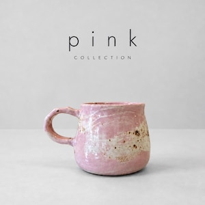 May include: A handmade ceramic mug with a rounded shape and a side handle. The surface features a mottled pink and white glaze with brown speckled accents. Above the mug, the text reads "p i n k COLLECTION" in black lettering.