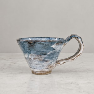 Peut inclure: Une tasse en céramique avec un bord large et évasé ainsi qu'une anse texturée et irrégulière. L'extérieur présente un émail moucheté dans des tons bleu ardoise, blanc et brun terreux, avec des coups de pinceau visibles et un aspect artisanal.