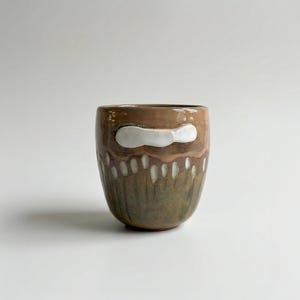 May include: A handmade ceramic cup with a brown glaze and white accents. The cup features a white, cloud-like shape near the top and vertical white dashes along the lower half. The cup is a unique piece of pottery.