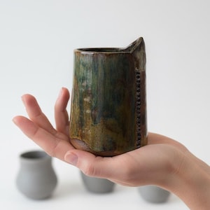 May include: A handcrafted ceramic vase with an unusual, asymmetrical shape. The vase, held in a hand, displays earthy green and brown hues. It has a textured surface and a cut-out detail on the rim. Other grey vases are in the background.
