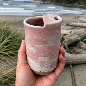 May include: A cylindrical ceramic travel mug with a pink and white marbled glaze finish. The mug features a built-in lid section with a small circular opening for drinking, and the base is unglazed, revealing the natural tan colour of the clay material.