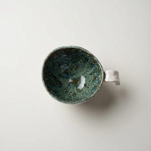 May include: A handmade ceramic mug with a speckled teal and green interior and a white handle. The mug has a unique, organic shape and a glossy finish. The handle is curved and attached to the side of the mug.