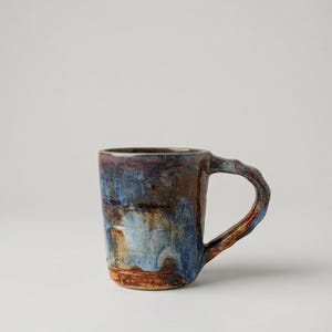 May include: A handcrafted ceramic mug with a distinctive, textured handle. The mug showcases a mix of blue, brown, and rust-toned glazes, producing a marbled appearance. The mug's design is simple yet artistic, ideal for tea or coffee.