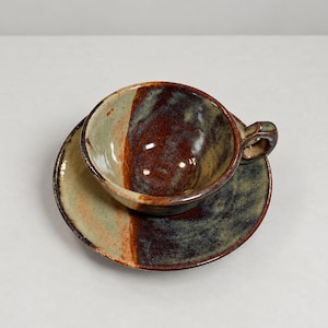 May include: A ceramic teacup and saucer set featuring a reactive glaze with a split color design. The surface displays a blend of earthy brown, deep rust, and muted sage green tones with a glossy finish. The cup has a small handle on the side.