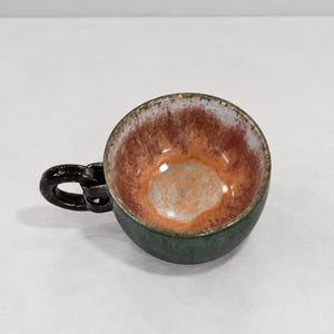May include: A ceramic cup with a dark green exterior and a glossy interior featuring a blend of orange, brown, and white glaze. The cup has a unique handle shaped like a dark brown chain link, providing a distinct visual contrast to the bowl.