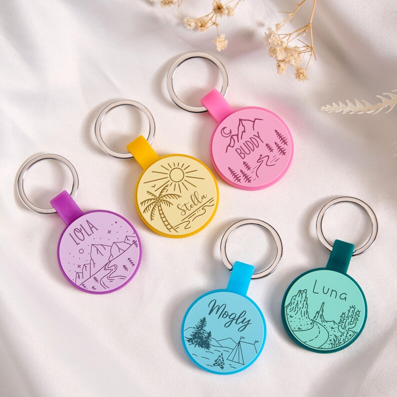 May include: A collection of colorful keychains with silver rings. Each keychain features a different color and design, including illustrations of mountains, palm trees, and camping scenes. The keychains have names like "Lola," "Stella," and "Buddy."