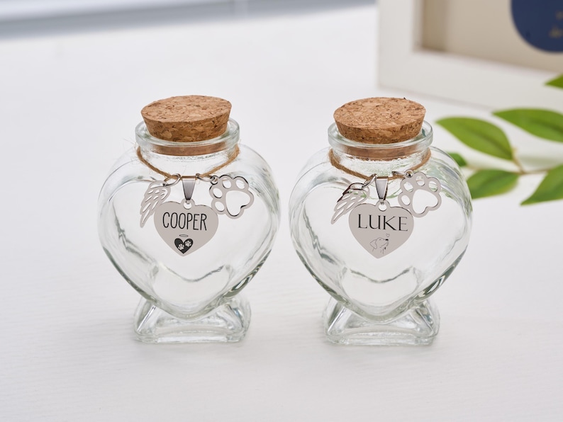 Peut inclure: Deux bocaux en verre transparent en forme de c&oelig;ur avec des bouchons en li&egrave;ge. Chaque bocal porte un charm argent&eacute; avec une empreinte de patte, une aile et un pendentif en forme de c&oelig;ur avec les noms des animaux de compagnie, "COOPER" et "LUKE".