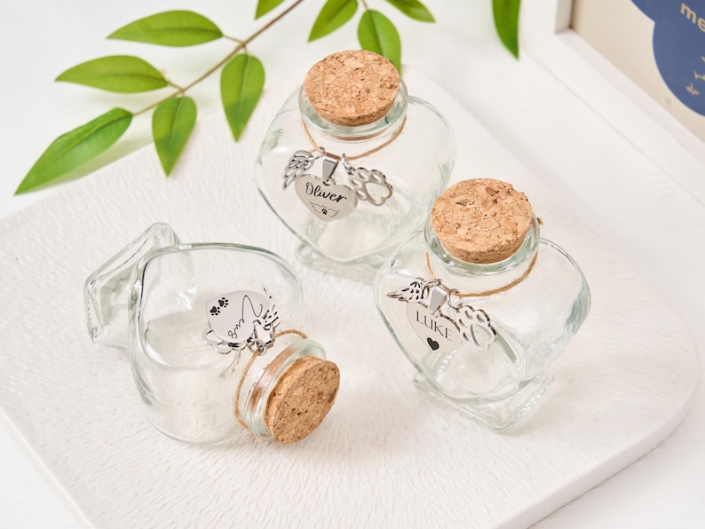 Peut inclure: Trois bouteilles en verre transparent en forme de c&oelig;ur avec des bouchons en li&egrave;ge. Chaque bouteille a un charm argent&eacute; avec un motif sur le th&egrave;me des animaux de compagnie et un nom : "Oliver" et "Luke". Les bouteilles sont pos&eacute;es sur un plateau blanc.