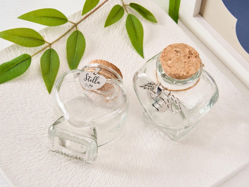 Peut inclure: Deux bouteilles en verre transparent en forme de c&oelig;ur avec des bouchons en li&egrave;ge. L'une porte une &eacute;tiquette &laquo; Stella &raquo;. L'autre a un charme en argent. Les bouteilles sont sur une surface blanche textur&eacute;e.