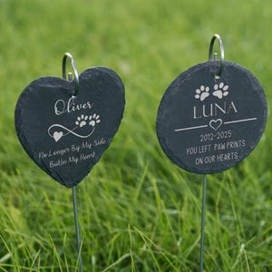 May include: Two dark gray slate pet memorial garden stakes. One is heart-shaped with the name "Oliver", paw prints, and the text "No Longer By My Side But In My Heart". The other is round with "LUNA", paw prints, and the text "You left paw prints on our hearts".