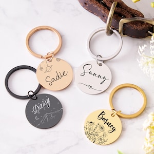 May include: A collection of personalized pet ID tags in various colors, including gold, silver, and black. Each tag is round and features a name and decorative elements like flowers, planets, or mountains. The tags are attached to key rings.