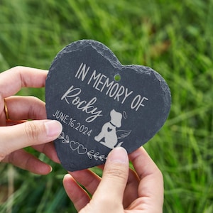 May include: A heart-shaped, dark gray slate memorial stone. The inscription reads "IN MEMORY OF Rocky JUNE.16.2024" with a dog illustration. The stone is held against a blurred green background.