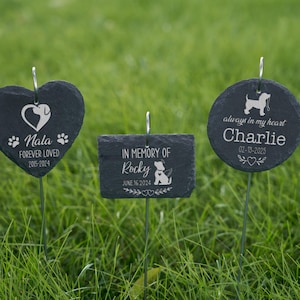 May include: Three black slate pet memorial markers in a grassy setting. One is heart-shaped, one rectangular, and one circular. Each has a pet's name, dates, and a dog silhouette. The heart-shaped marker says "Nala Forever Loved 2015-2024". The rectangular marker says "In Memory of Rocky June 16 2024". The circular marker says "always in my heart Charlie 02-13-2025".