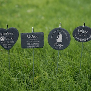 May include: Four dark gray slate pet memorial markers with silver metal stakes. The markers are heart, rectangular, round, and oval shaped. Each marker has a name and dates engraved, with phrases like "In Memory Of" and "Forever Missed". They are set in green grass.
