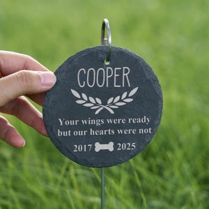 May include: A round, dark gray slate pet memorial marker with the name "COOPER" in white script. The marker includes a laurel wreath design and the text "Your wings were ready but our hearts were not" with the years 2017 and 2025.