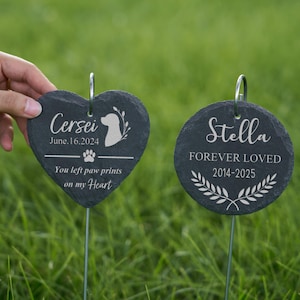 May include: Two dark gray slate pet memorial garden stakes. One is heart-shaped with the name "Cersei", date, paw print, and text. The other is round with the name "Stella", dates, and text. Both have metal stakes.