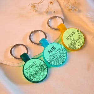 Può includere: Tre portachiavi personalizzati con anelli argentati. Ogni portachiavi ha una base rotonda e colorata con un nome e un'illustrazione a tema natura. I portachiavi sono verdi, blu e gialli. I nomi sono "Tucker", "Alfie" e "Maggie".