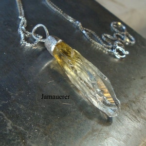 May include: A silver chain necklace with a clear quartz crystal pendant. The crystal is a long, thin point with a yellow hue.