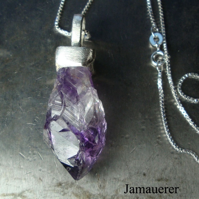 May include: A silver chain necklace with a pendant featuring a large, faceted amethyst crystal. The crystal is a deep purple color and has a pointed shape.