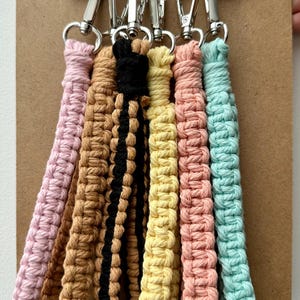 Peut inclure: Sept porte-clés en macramé colorés avec des fermoirs métalliques accrochés à un presse-papiers. Les porte-clés sont dans des tons de rose, marron, noir, jaune et bleu.