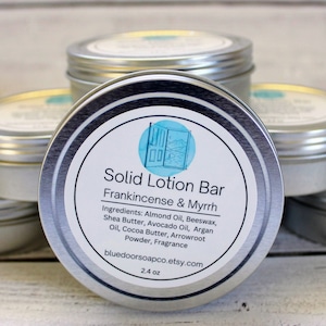 May include: A silver tin with a white label that reads "Solid Lotion Bar Frankincense & Myrrh". The label also lists the ingredients: Almond Oil, Beeswax, Shea Butter, Avocado Oil, Argan Oil, Cocoa Butter, Arrowroot Powder, and Fragrance. The tin is 2.4 oz and is made by bluedoorsoapco.etsy.com.