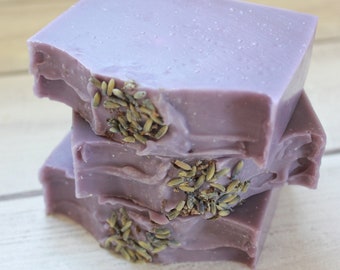 May include: Three bars of lavender soap stacked on top of each other. The soap is a light purple color and has dried lavender buds embedded in it.