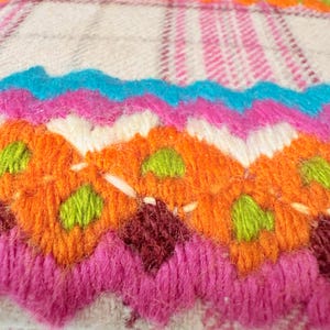 May include: A colorful, hand-stitched textile with a geometric design. The piece features vibrant hues of pink, orange, blue, and green, creating a striking visual contrast. The texture appears soft and plush, suggesting a cozy, handcrafted item.