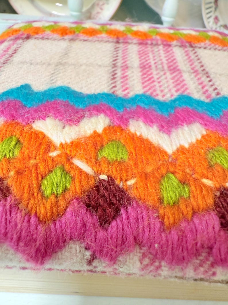 May include: A colorful, hand-stitched textile with a geometric design. The piece features vibrant hues of pink, orange, blue, and green, creating a striking visual contrast. The texture appears soft and plush, suggesting a cozy, handcrafted item.