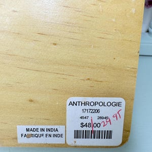 May include: A light-colored wooden item with visible wood grain. Two stickers are attached: one reads "Made in India / Fabriqu&eacute; en Inde," and the other displays "ANTHROPOLOGIE" and a barcode.