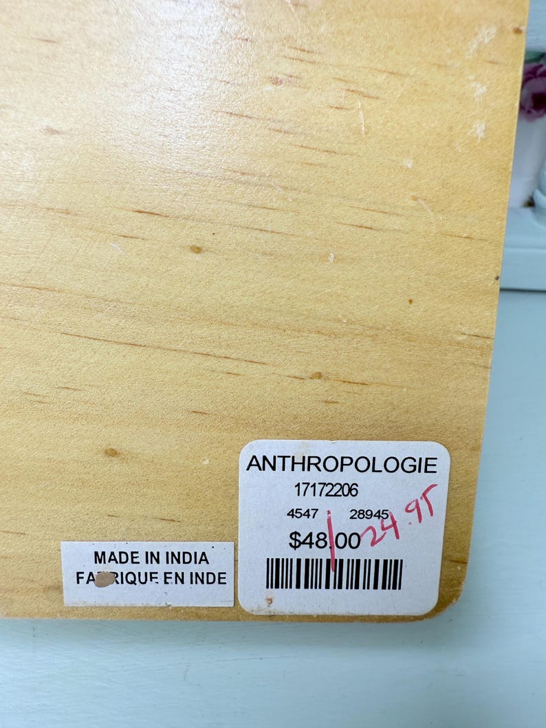 May include: A light-colored wooden item with visible wood grain. Two stickers are attached: one reads "Made in India / Fabriqu&eacute; en Inde," and the other displays "ANTHROPOLOGIE" and a barcode.