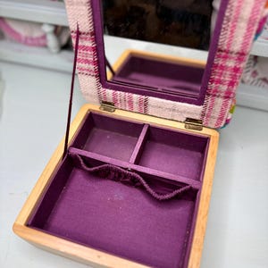 May include: Open wooden jewelry box with a purple interior and a plaid fabric lid. The lid has a built-in mirror. The box is open, revealing compartments for storing jewelry. The fabric on the lid is pink, white, and purple.